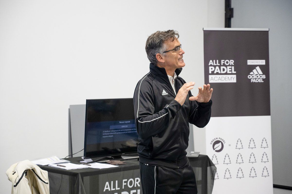 Un uomo parla durante una presentazione dell'accademia di padel, indossa una tuta nera Adidas e gesticola con le mani.