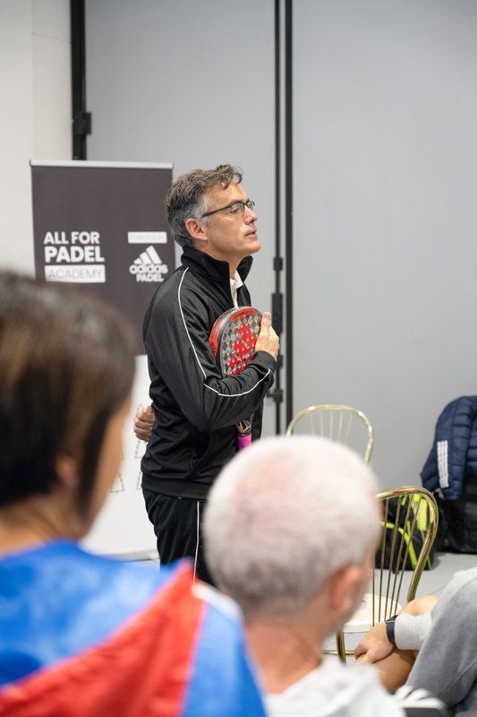 Un uomo con la racchetta parla di fronte a persone sedute; ambiente interno, marchio Adidas sullo sfondo.