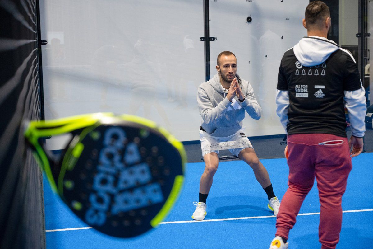 Uomo che fa stretching sul campo da padel, l'allenatore lo osserva; racchetta in primo piano.
