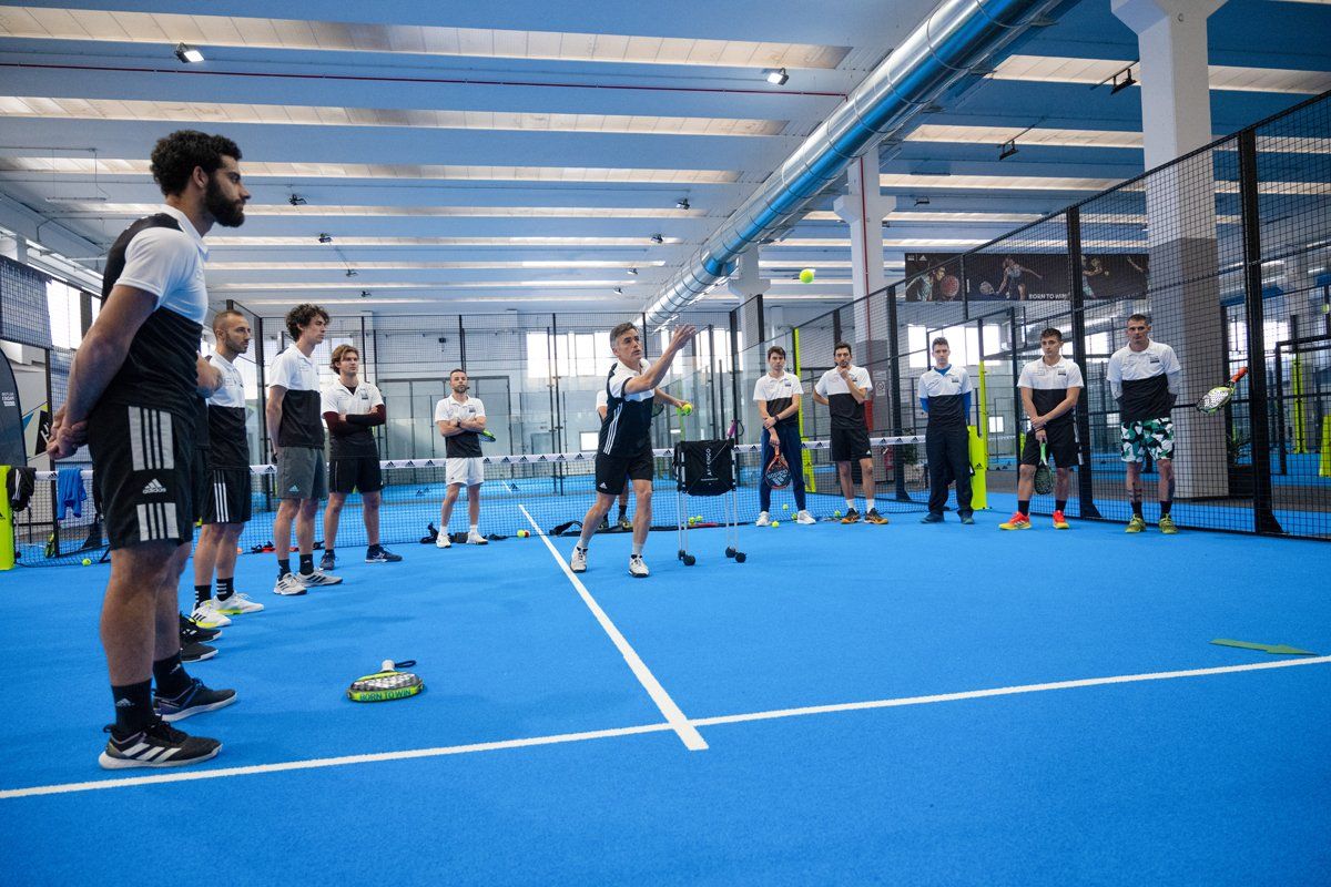 Un gruppo di persone su un campo da padel blu osserva un giocatore che dimostra un servizio, all'interno di una palestra