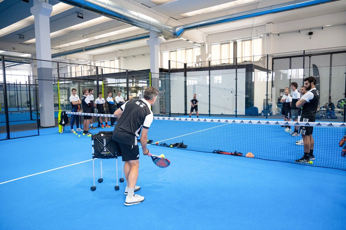Lezione di padel tennis al chiuso: l'allenatore mostra il gioco, gli studenti guardano, campo blu, rete e attrezzatura.