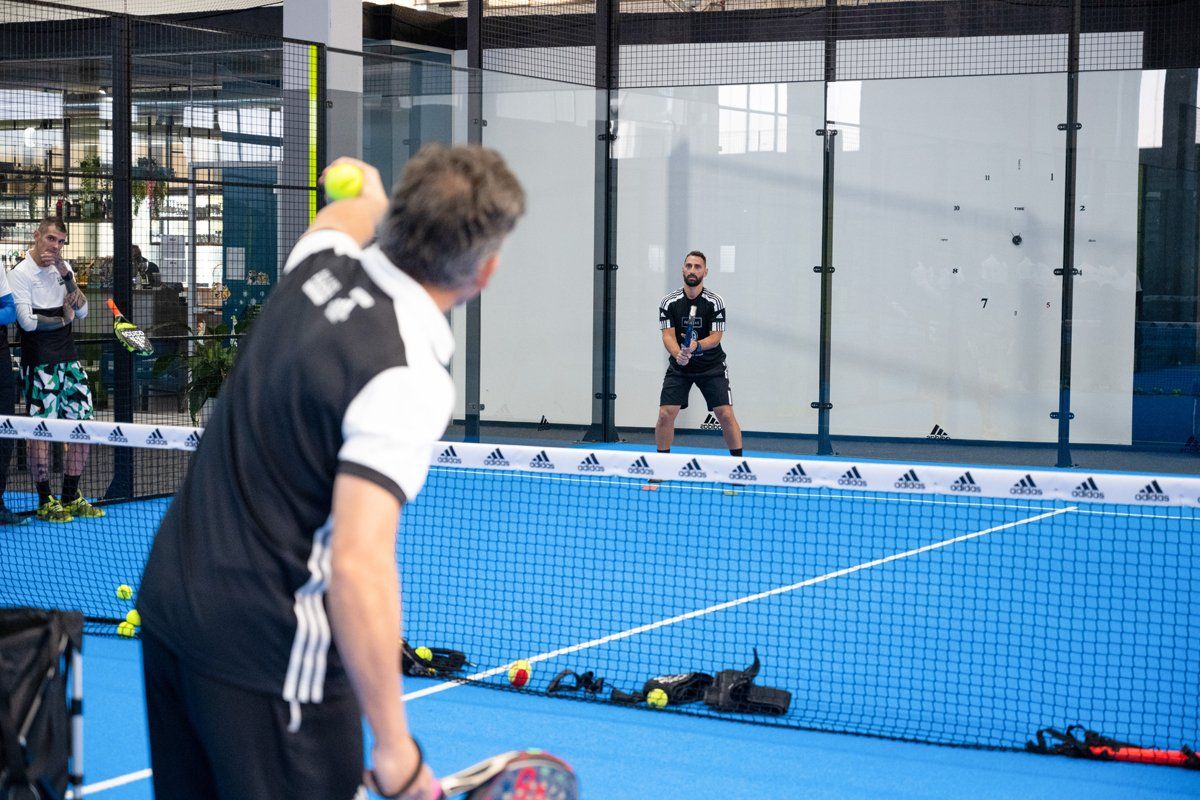 Un uomo serve una pallina gialla da paddle tennis sopra la rete, un altro giocatore è pronto a riceverla su un campo blu.