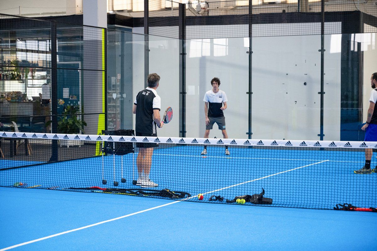 Tre persone giocano a padel tennis su un campo blu con rete; ambiente al coperto.