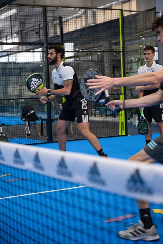 Persone che giocano a padel, un uomo serve, altri guardano. Campo con superficie blu, rete in primo piano.