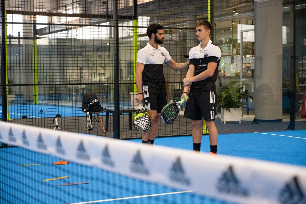 Due uomini su un campo da padel. Uno offre incoraggiamento, entrambi con le racchette in mano, con la rete in primo piano.