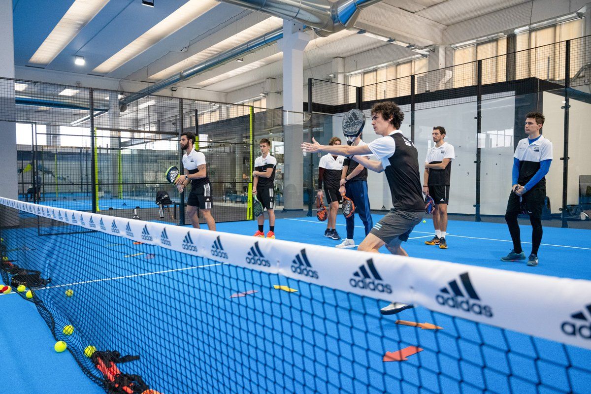 Persone che giocano a padel in un campo coperto con pavimento blu e rete, concentrandosi sul servizio e sul volée.