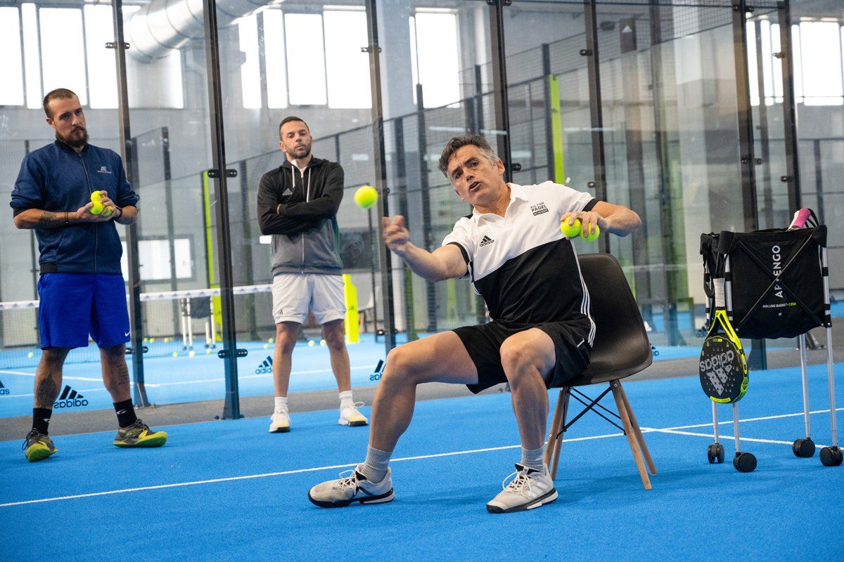 Tre uomini in un campo da padel. L'uomo al centro lancia una palla mentre gli altri osservano. Campo blu, ambiente neutro.