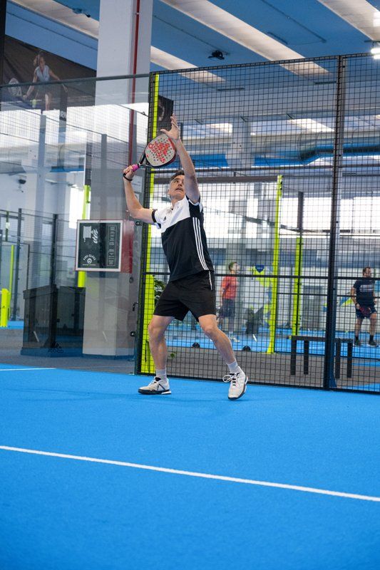Uomo che gioca a padel, alza la racchetta per un tiro dall'alto su un campo blu, al coperto.