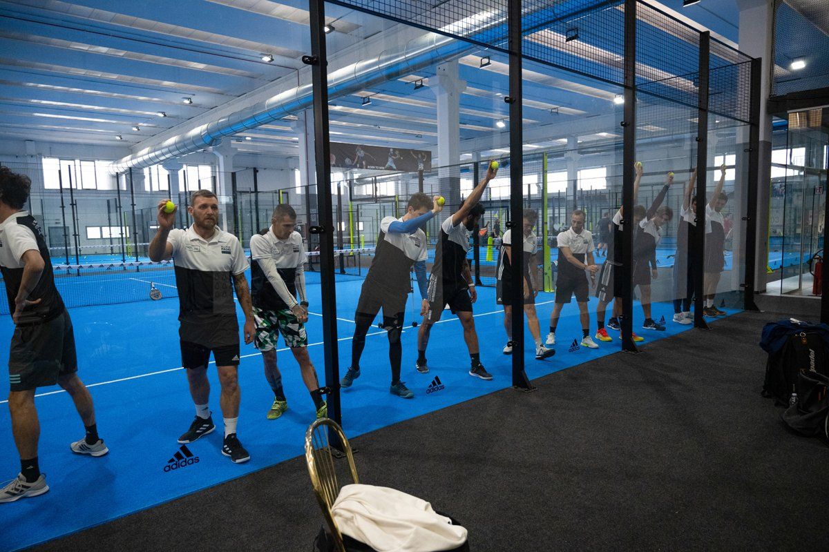 Persone che si allenano in un campo da padel al coperto, con palline e racchette in mano, campo blu, pareti grigie.