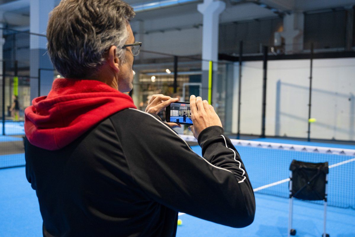 Uomo che filma un campo da paddle tennis con lo smartphone. Indossa una giacca nera con cappuccio rosso e occhiali.