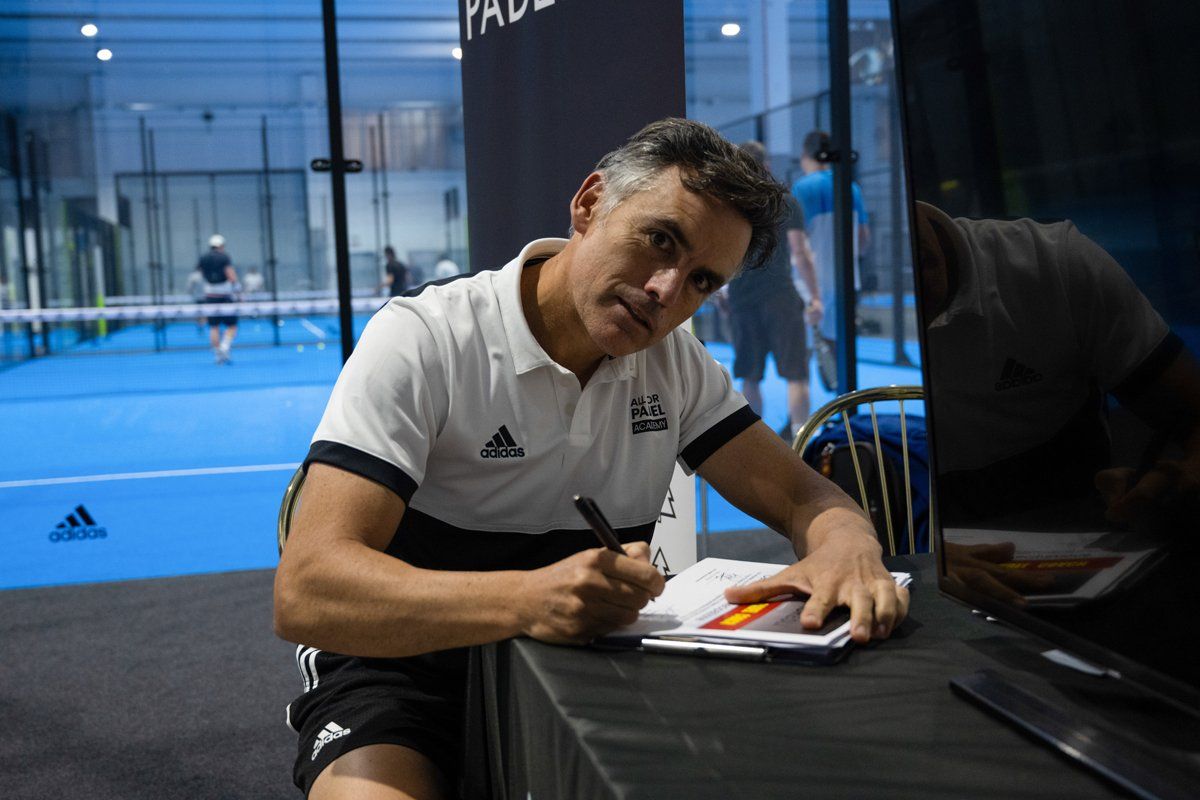 Un uomo firma autografi su un campo da padel. Indossa una polo e pantaloncini, con una bandiera ben visibile.