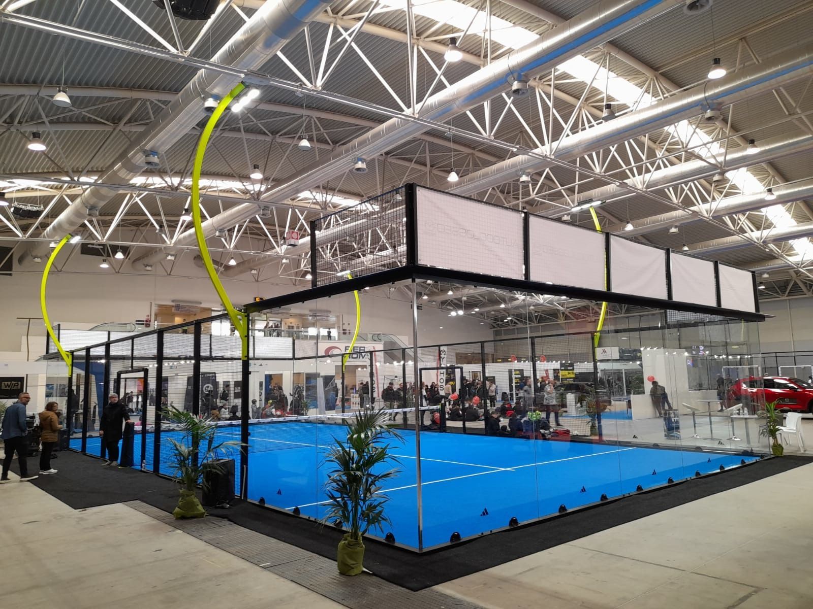 Campo da padel indoor con superficie blu e recinzione nera, persone che guardano.