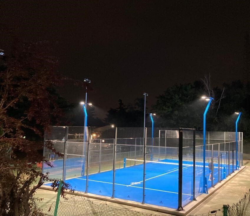 Campo da padel illuminato di blu di notte, circondato da alberi e alti lampioni.