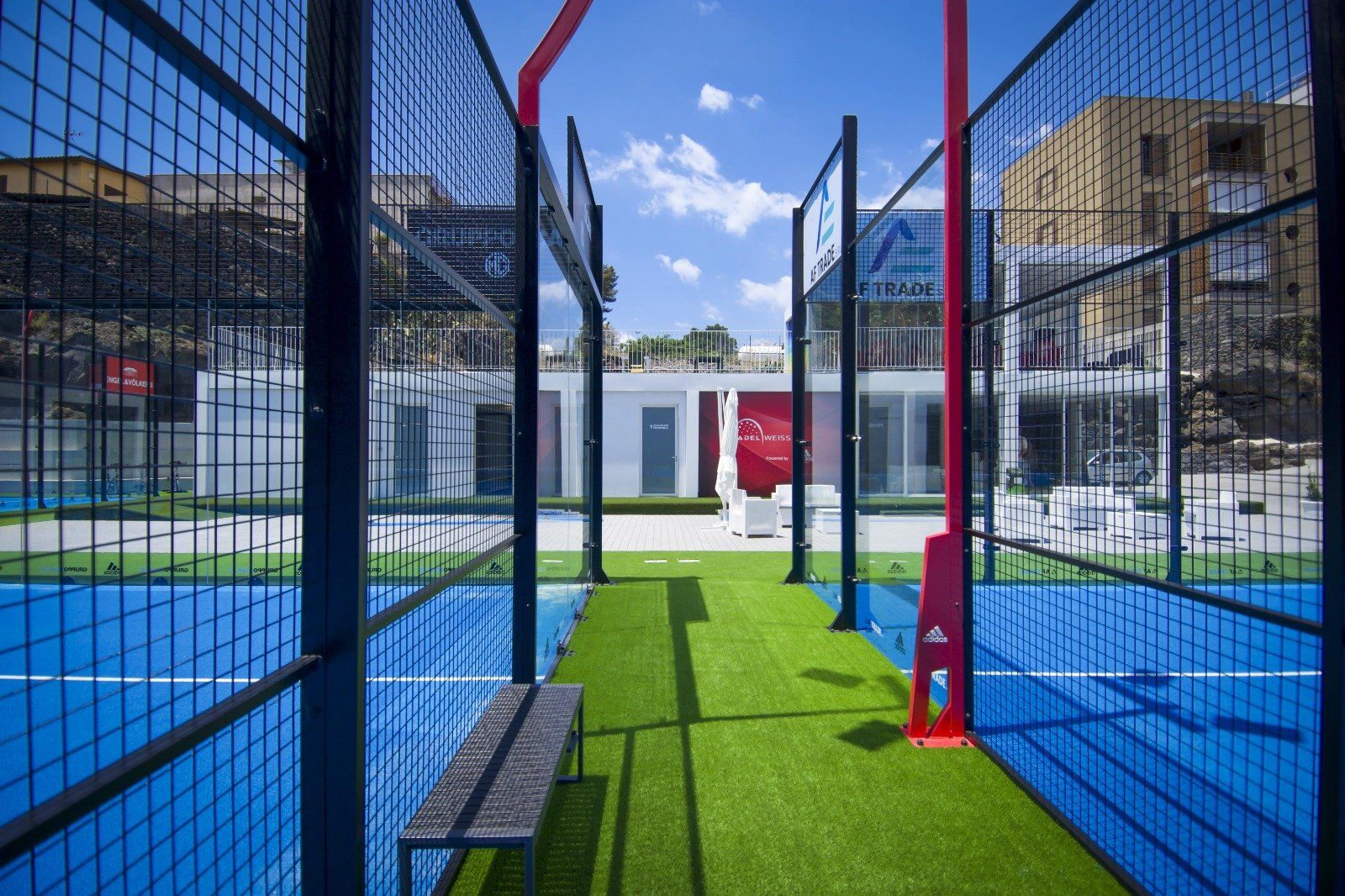 Un campo da padel blu con erba sintetica verde, una panchina nera e un palo rosso. Sullo sfondo un edificio bianco.