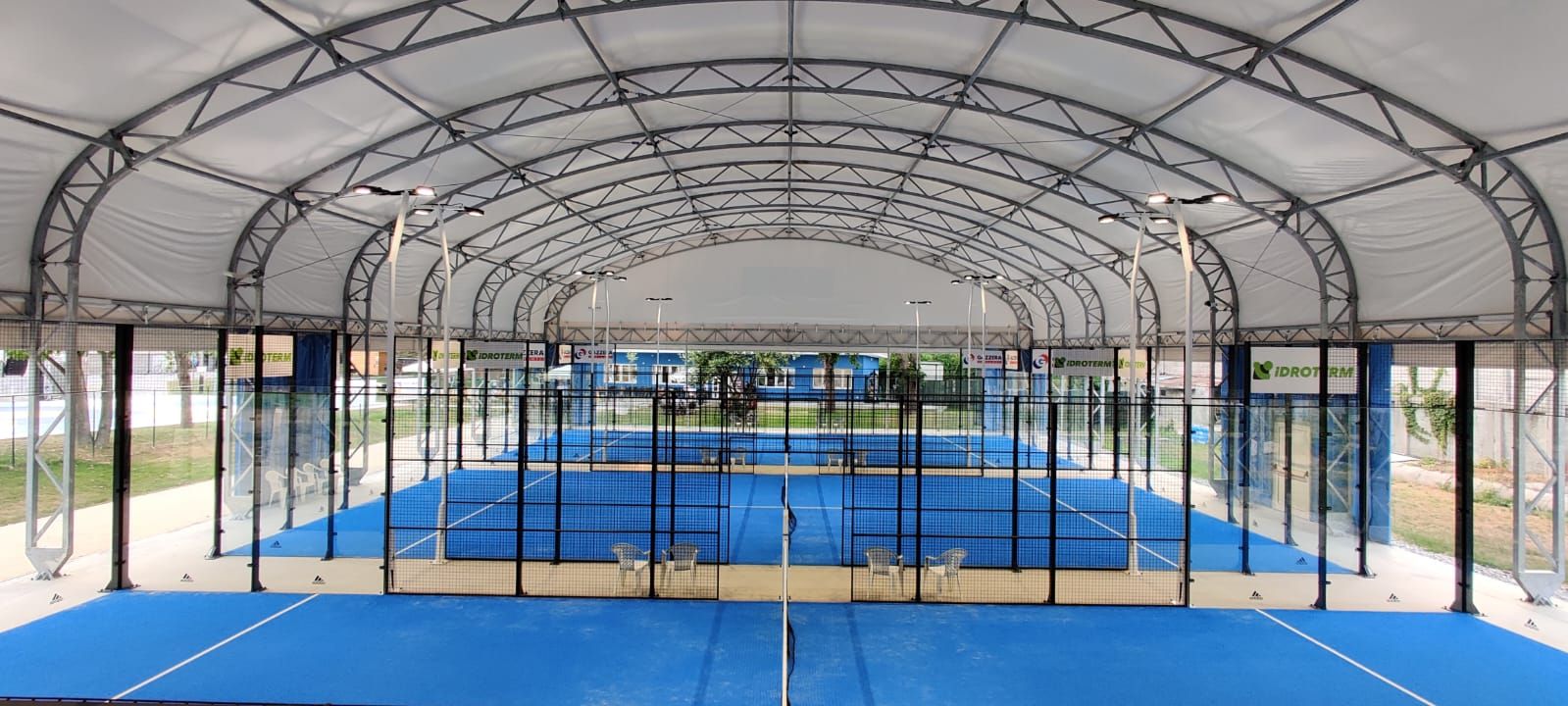 Campo da padel al coperto, superficie blu, pareti in vetro, tetto in metallo.