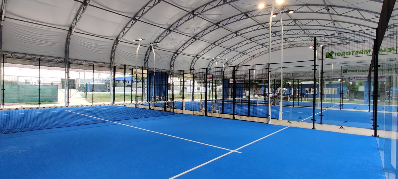 Campo da padel blu al coperto con pareti in vetro, linee bianche e tetto curvo.