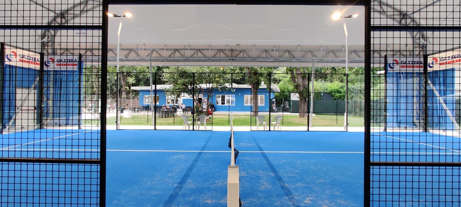 Campo da padel blu con rete, circondato da recinzioni e con un edificio in lontananza.