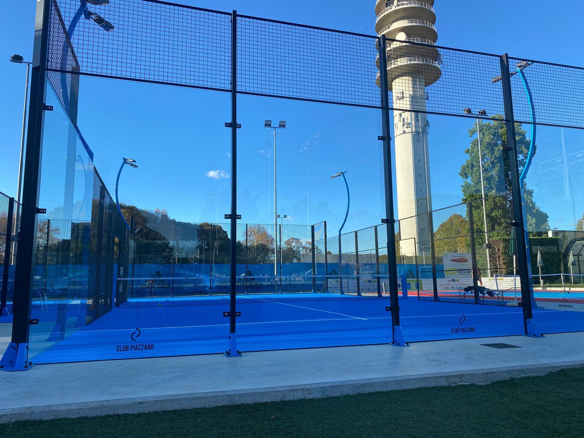 Campo da padel blu con pareti in vetro, rete nera e un'alta torre sullo sfondo in una giornata di sole.