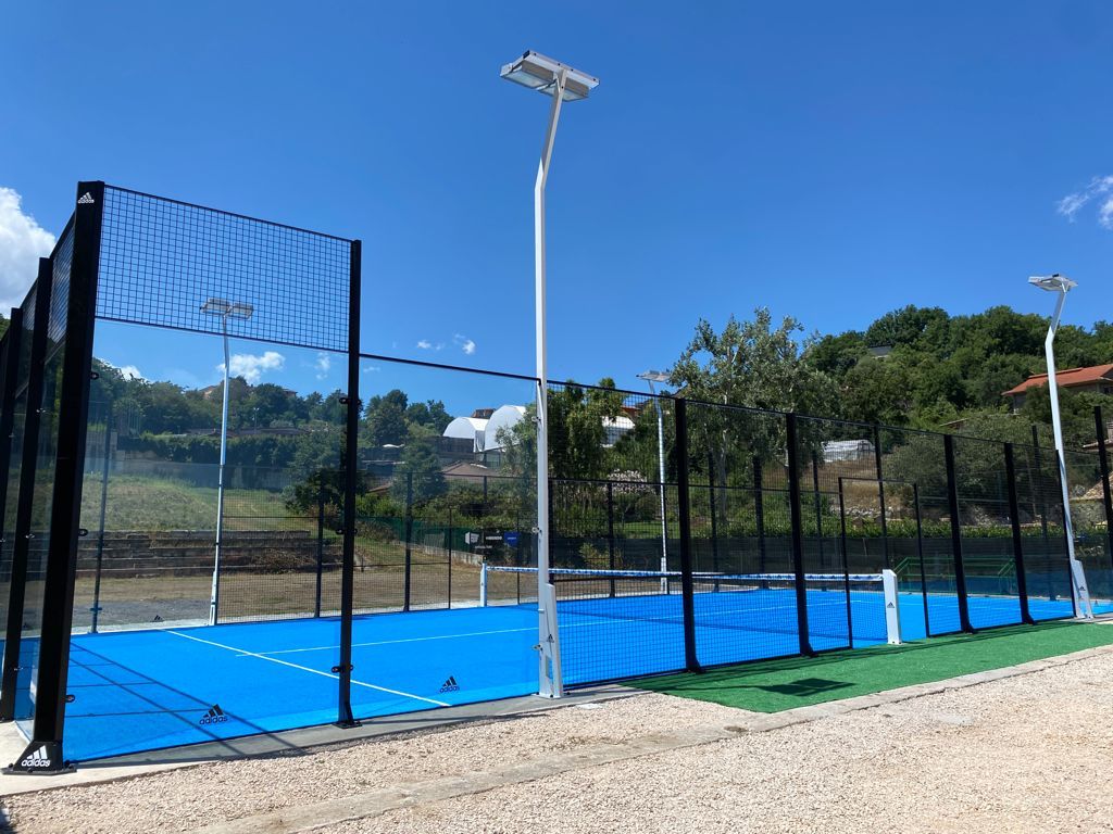 Campo da padel blu e verde, con rete nera, sullo sfondo di un cielo azzurro e soleggiato e di colline.