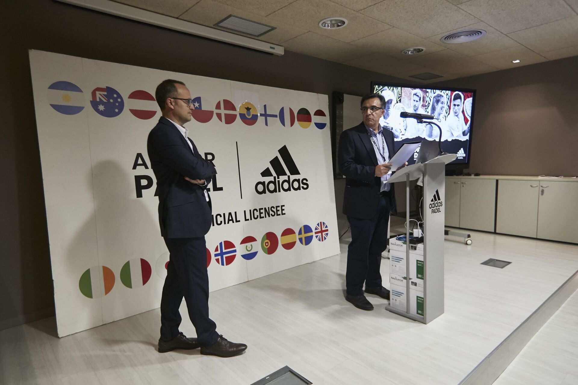Due uomini su un podio durante una presentazione. Sullo sfondo il logo Adidas e le bandiere. Un uomo parla.