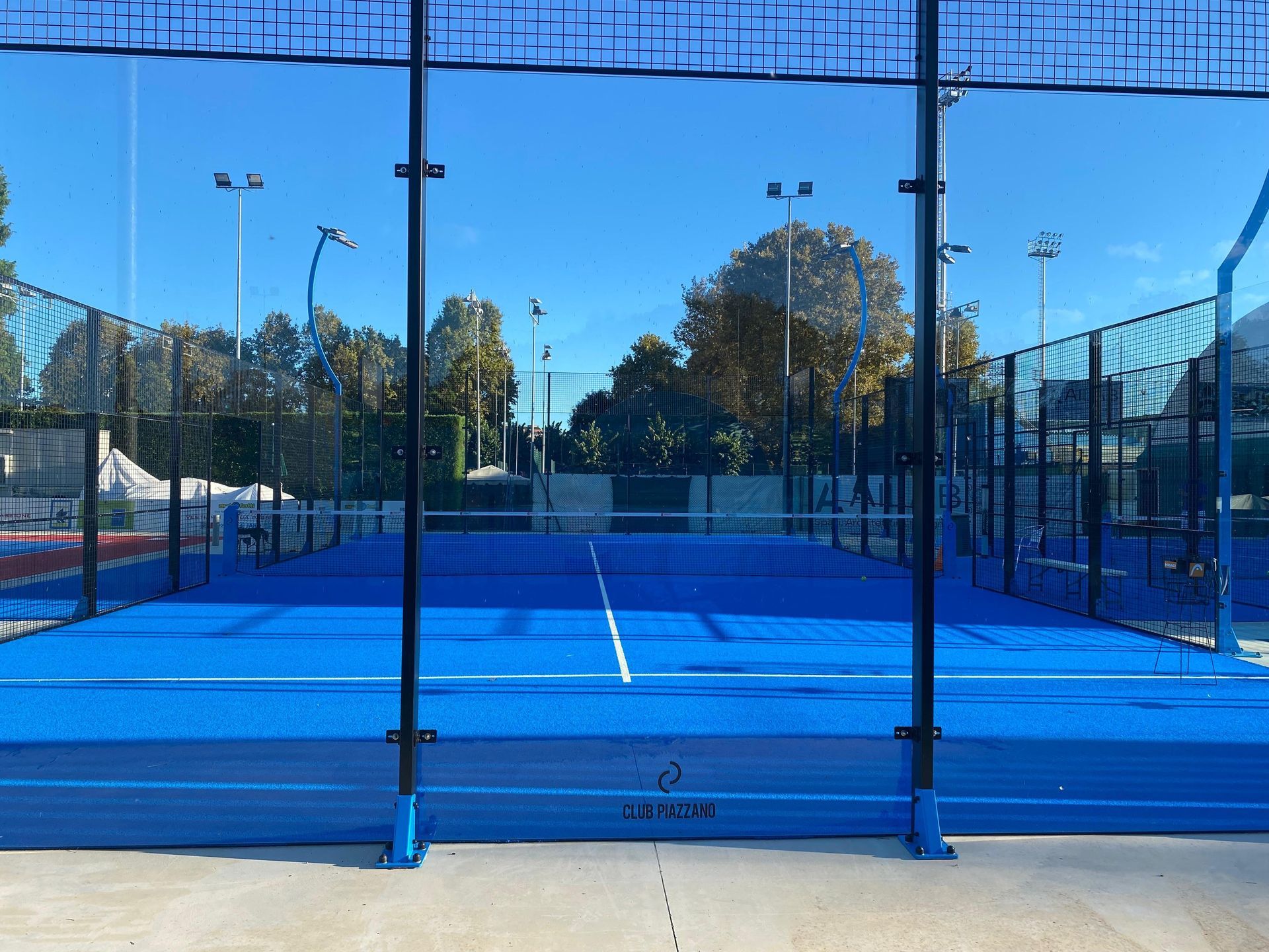Campo da padel blu con rete, circondato da pareti di vetro, sotto un cielo azzurro e terso.