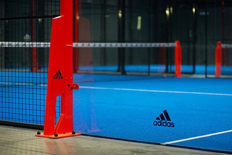Pilone arancione e logo Adidas su un campo da padel blu con linee bianche e una rete.