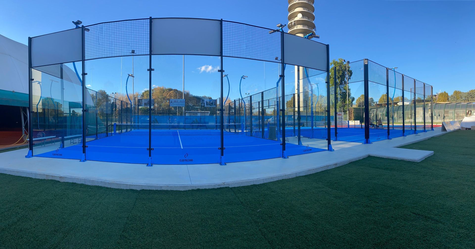 Campi da padel blu con pareti in vetro e superficie blu, circondati da erba artificiale verde.