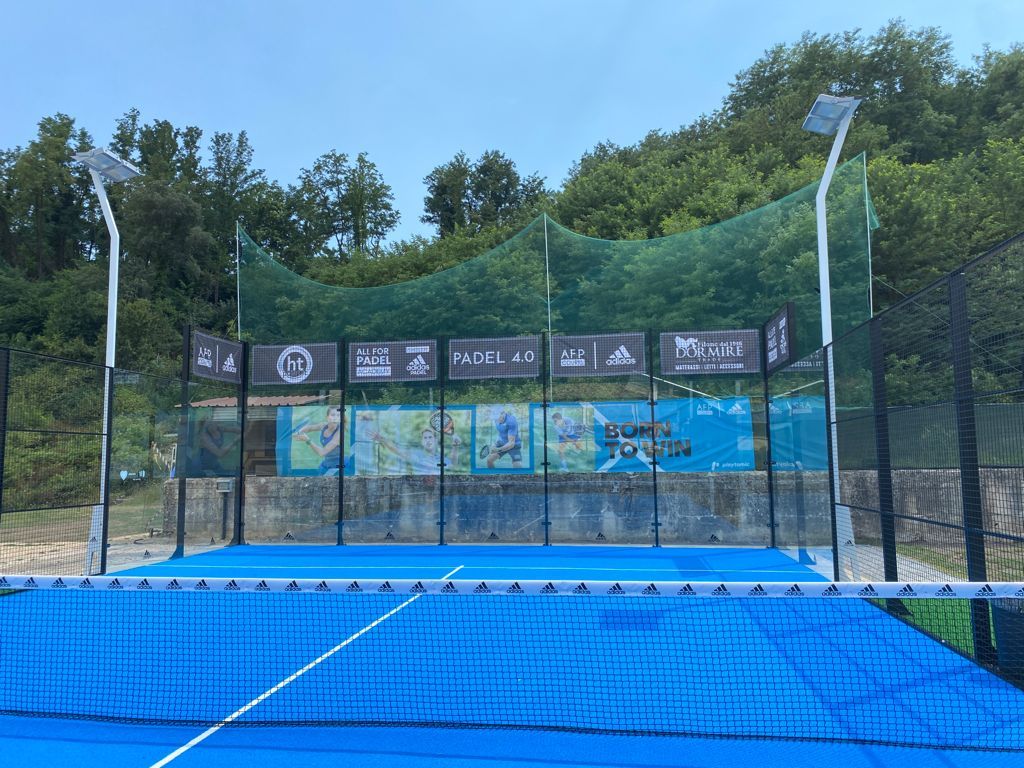 Campo da padel blu con rete, circondato da una recinzione verde, con striscioni degli sponsor e uno sfondo boschivo.