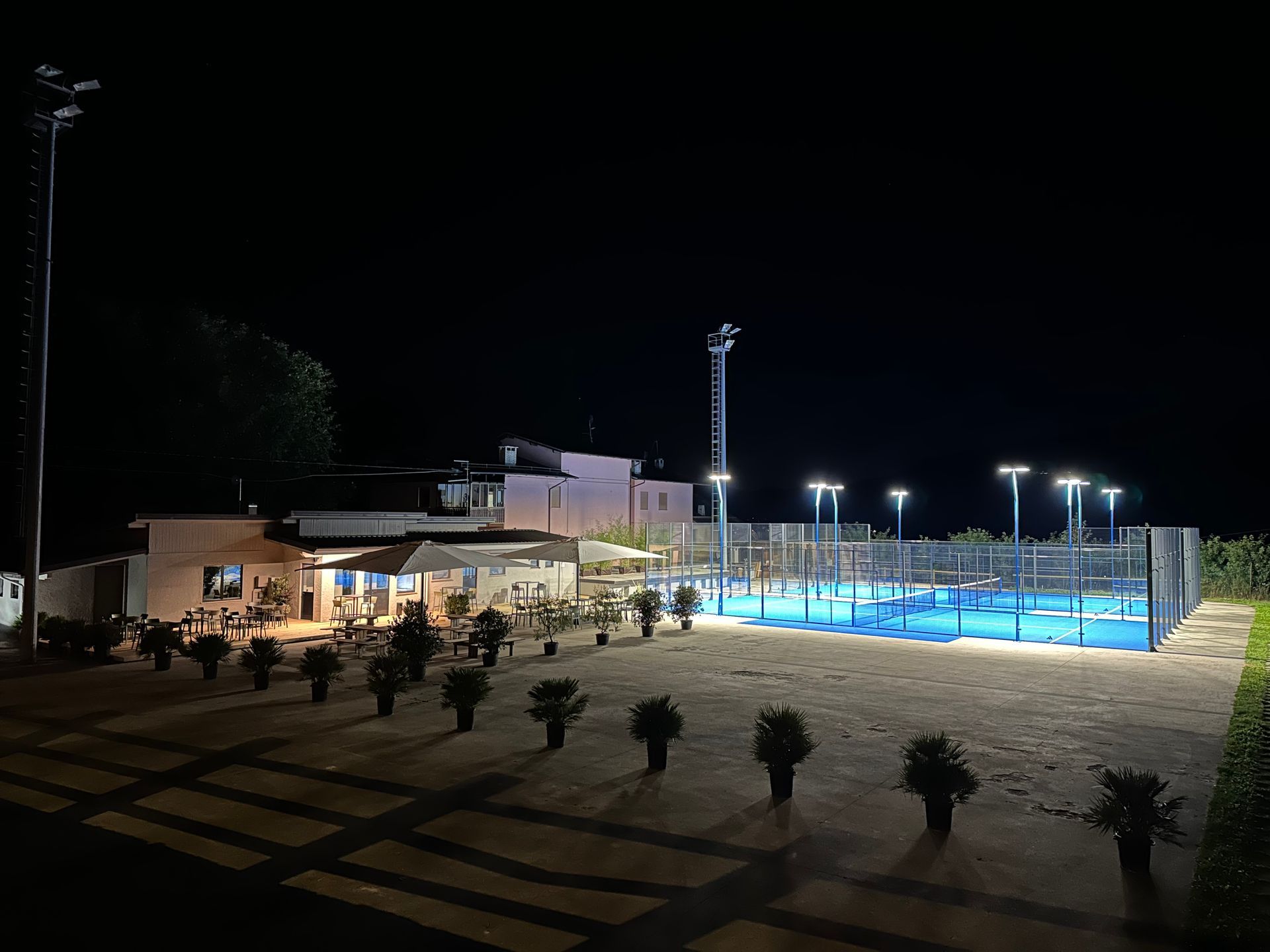 Vista notturna di un campo da padel ben illuminato, con un edificio e cespugli. Il campo ha superfici blu.