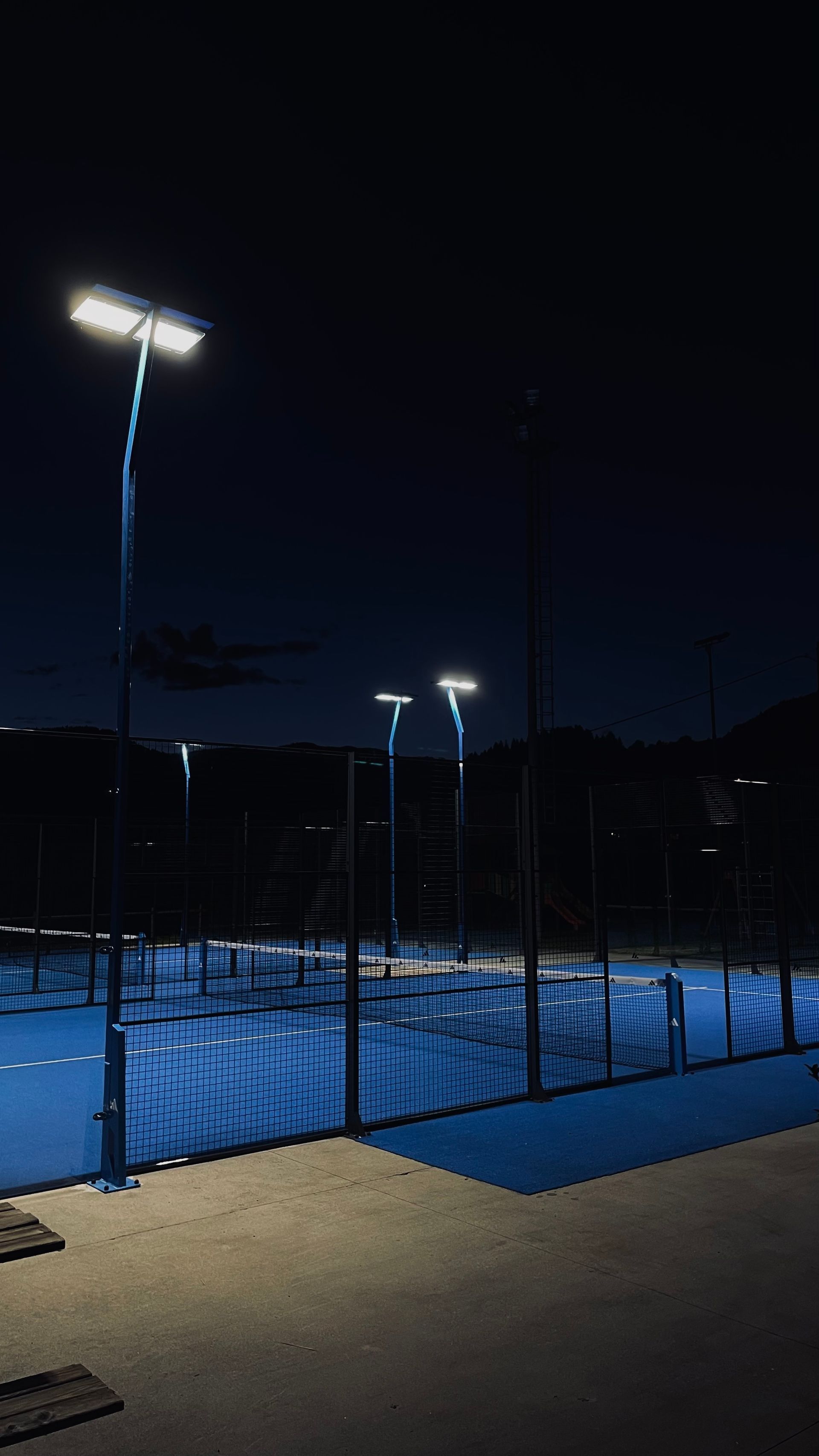 Vista notturna di un campo da padel illuminato da alte luci contro un cielo blu scuro. Un aereo vola sopra di noi.