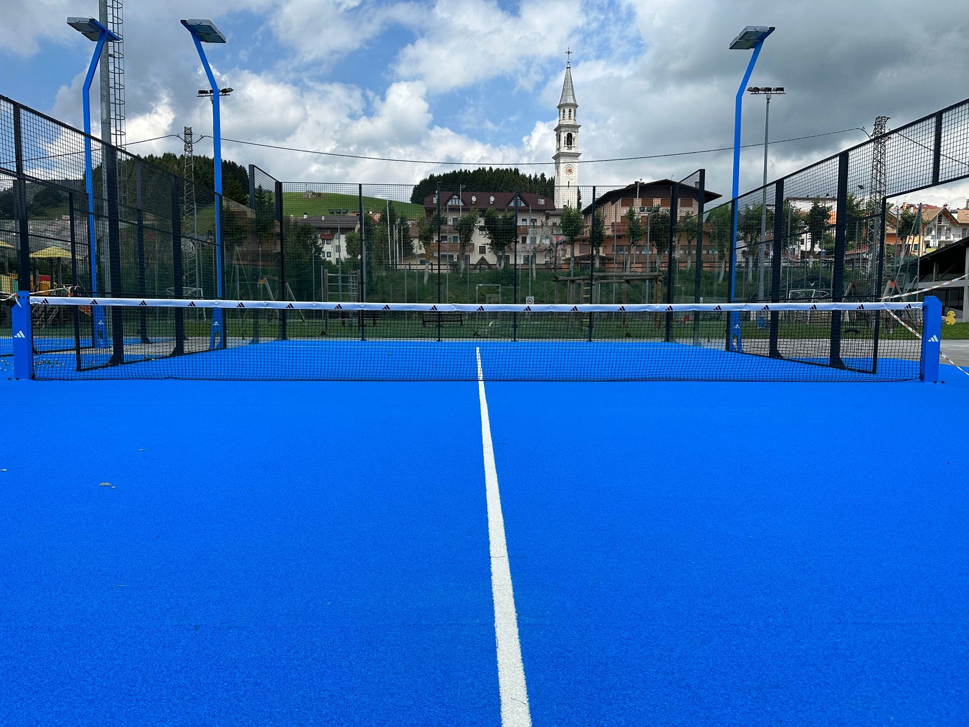 Campo da padel blu con una linea bianca, una rete e una città sullo sfondo sotto un cielo nuvoloso.