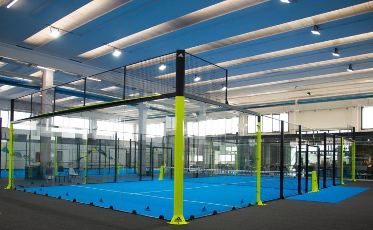 Campo da padel al coperto con pavimento blu, colonne di supporto gialle, rete e struttura nera.