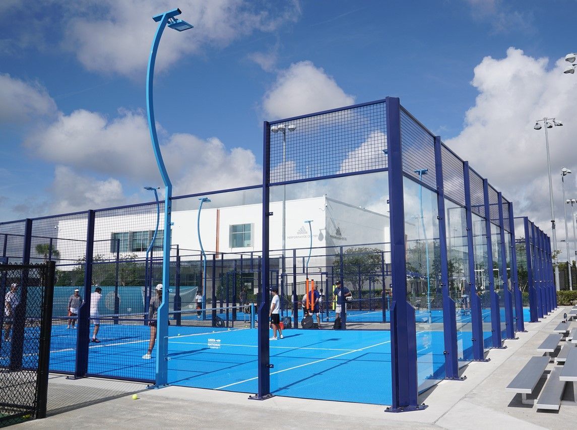 Campo da padel blu con giocatori, rete blu e nera, sotto un cielo luminoso.