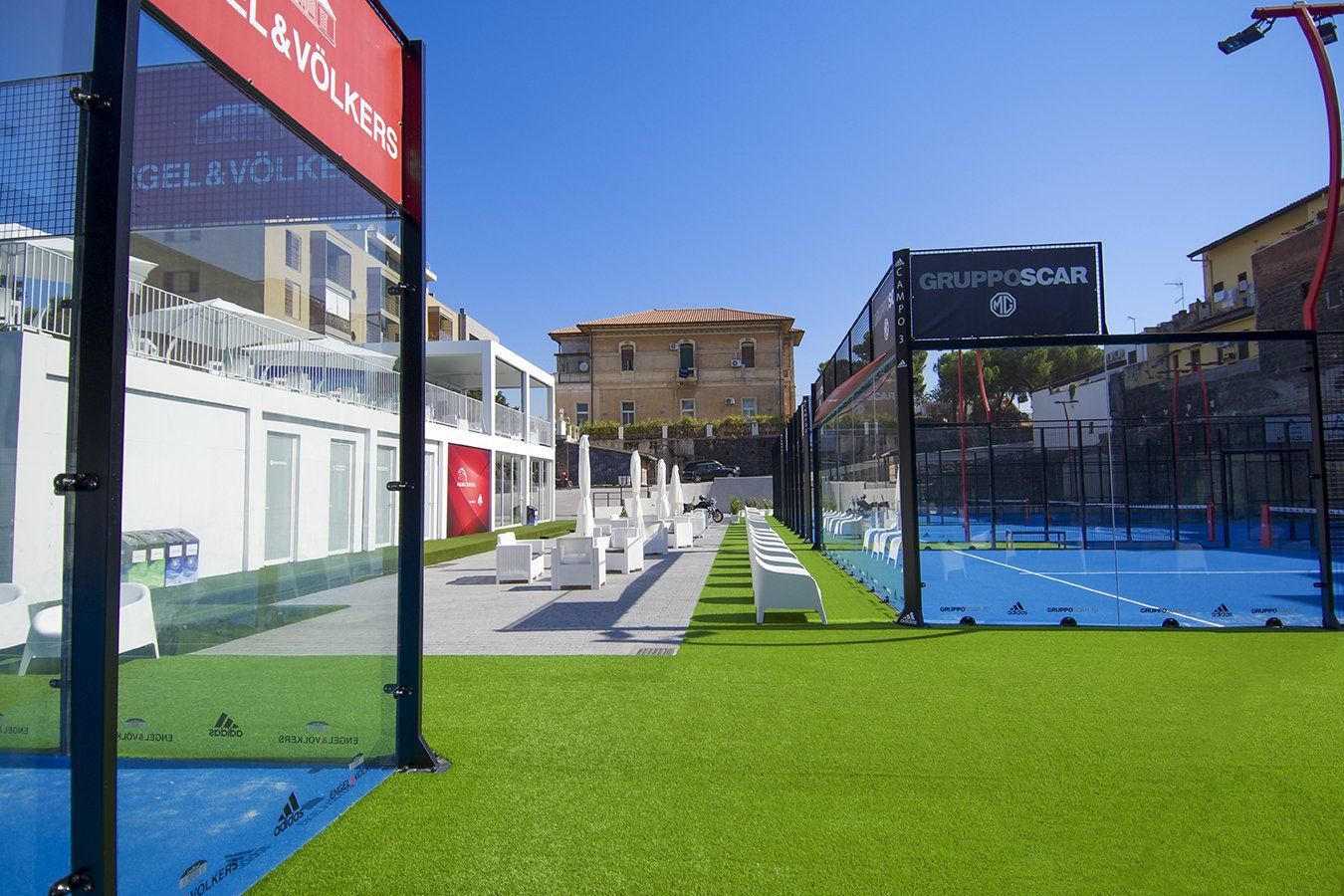 Campo da padel con superficie di gioco blu e manto erboso artificiale verde, accanto a un edificio bianco 