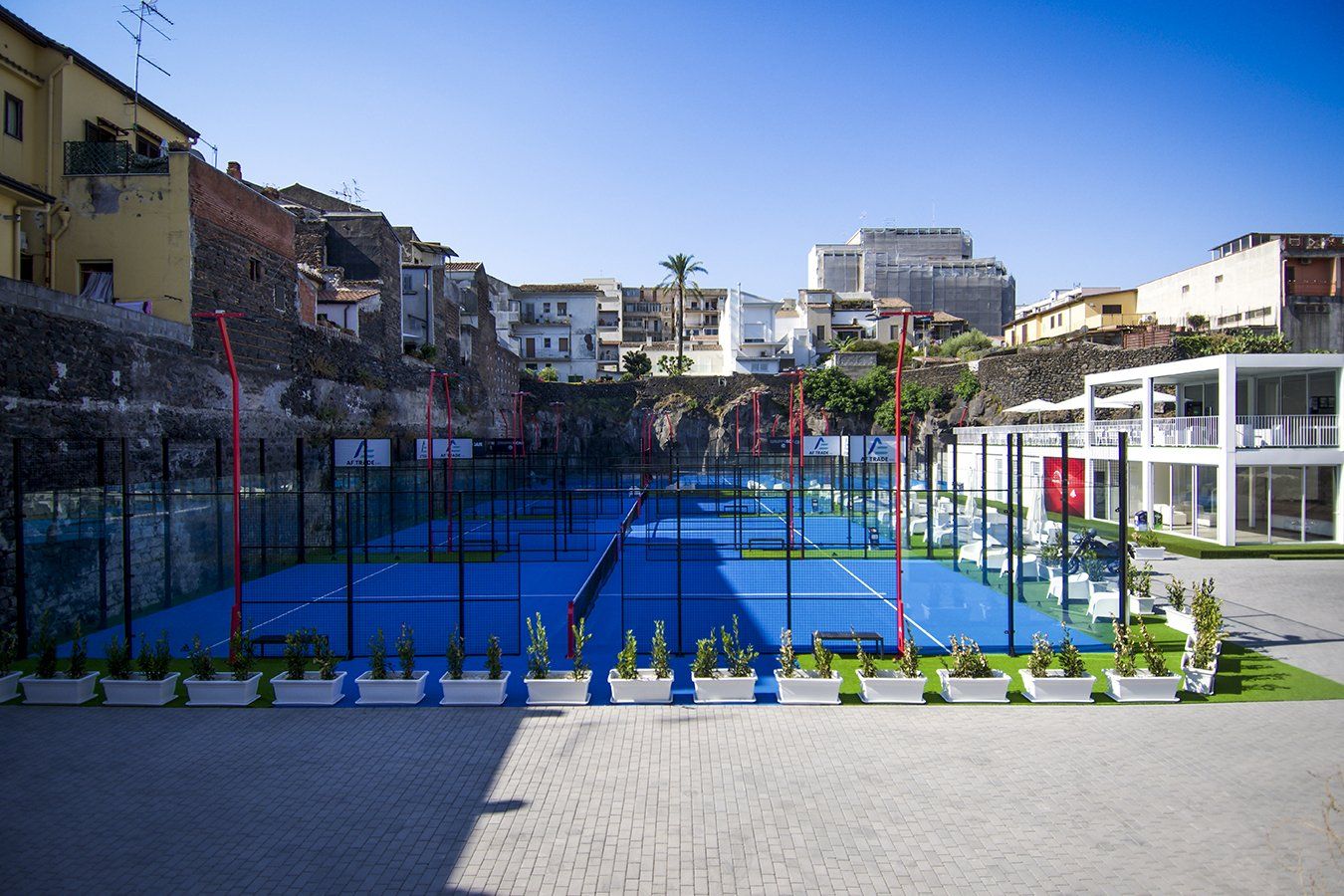 Campi da paddle blu con rete nera e pali rossi, sullo sfondo di un paesaggio urbano e di edifici.