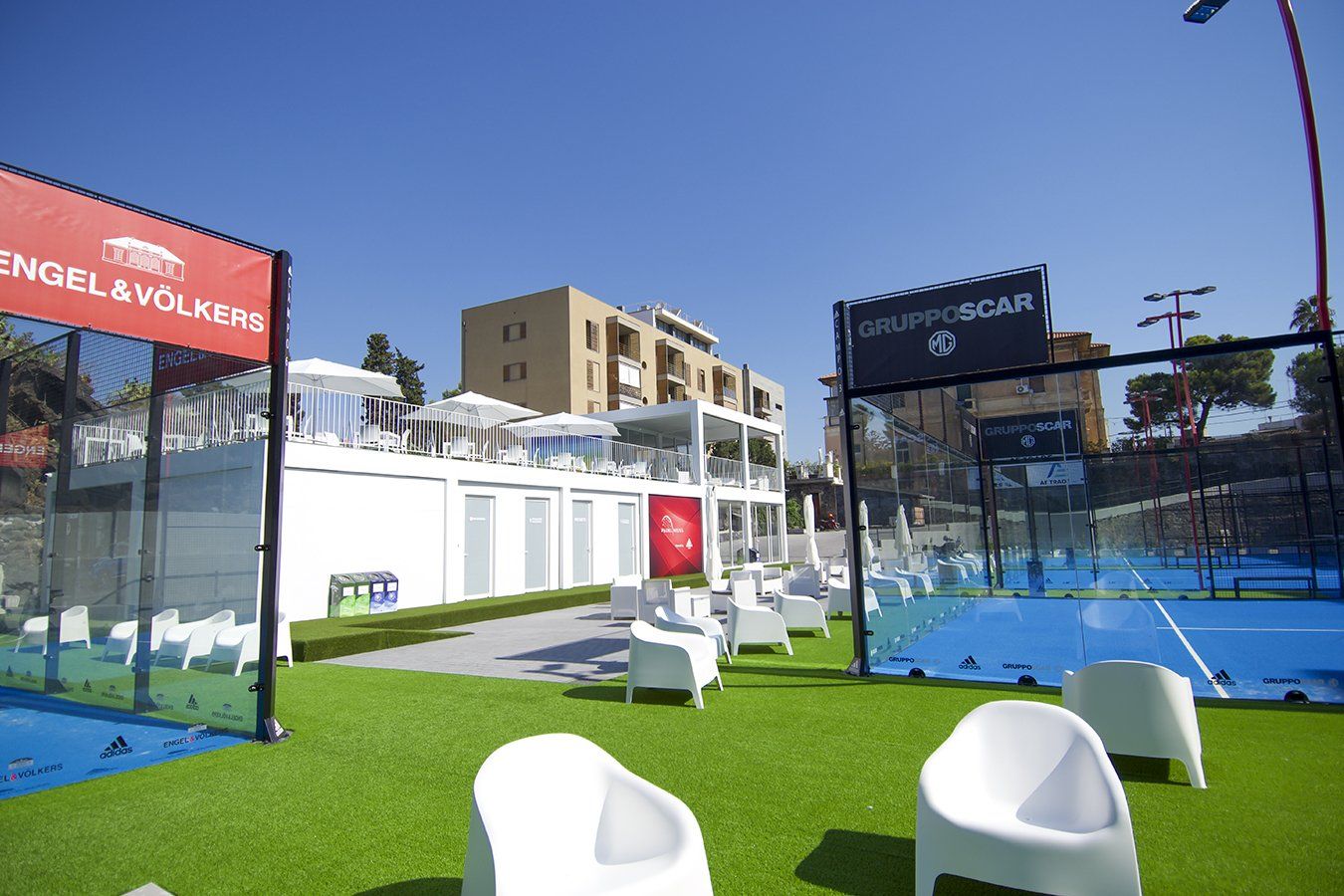 Campo da padel con superficie blu, erba artificiale verde, sedie bianche e un edificio sotto un cielo azzurro.