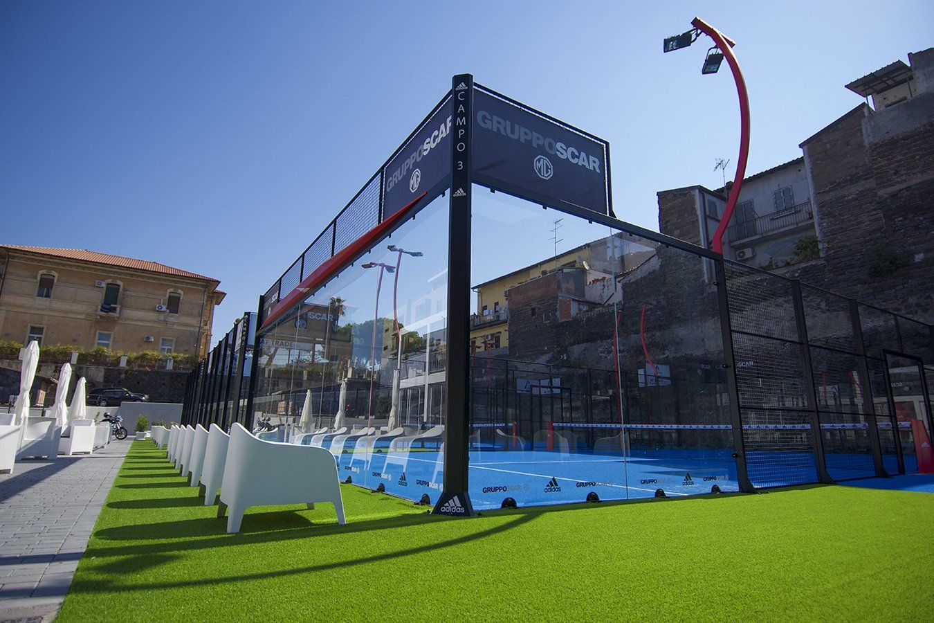 Campo da padel tennis in erba sintetica, circondato da pareti di vetro e recinzione metallica, in una giornata di sole.
