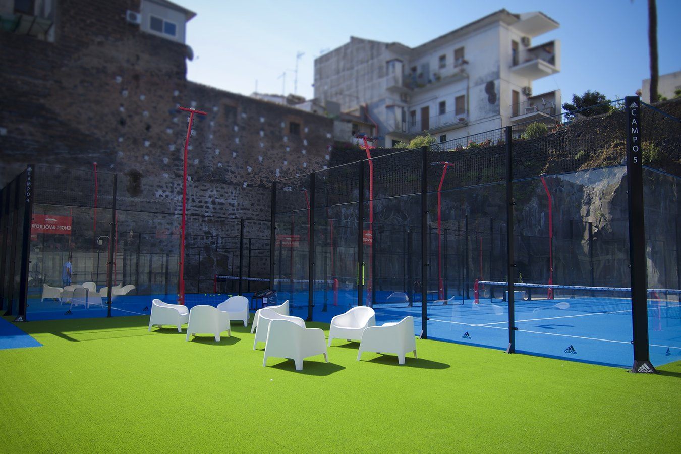 Campo da padel con erba verde, campo blu, sedie bianche e un edificio sullo sfondo.