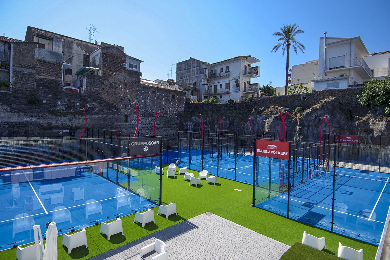 Campi da padel tennis all'aperto con superfici blu, erba verde e sedute bianche, sullo sfondo di edifici