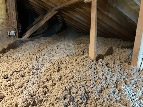 Attic filled with loose, light-colored insulation between wooden rafters and supports.