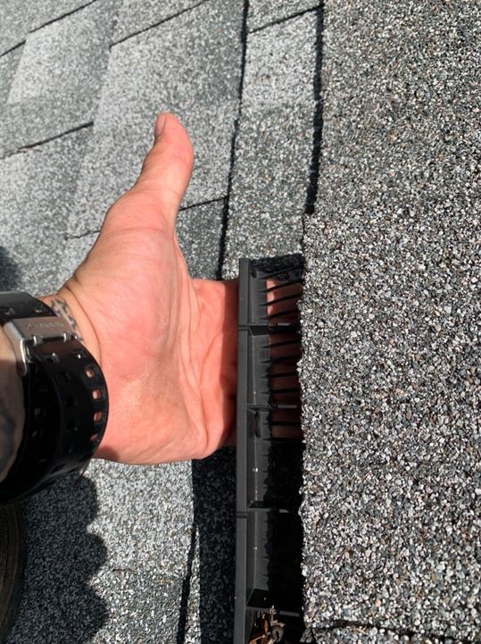Hand giving thumbs up next to roofing edge with a black strip. Gray shingles are visible.