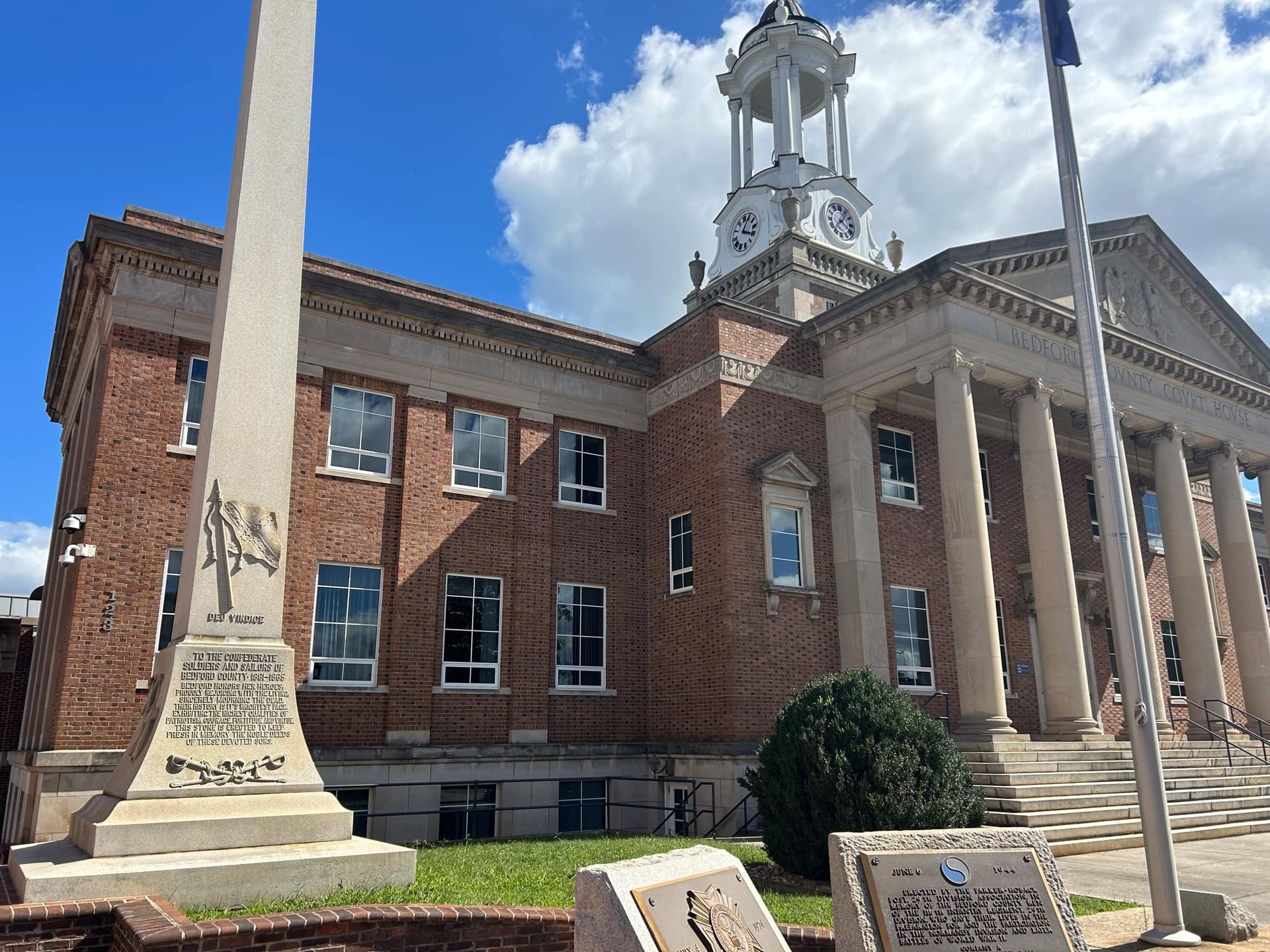 Brick courthouse with white columns, clock tower, and an obelisk monument on a sunny day.