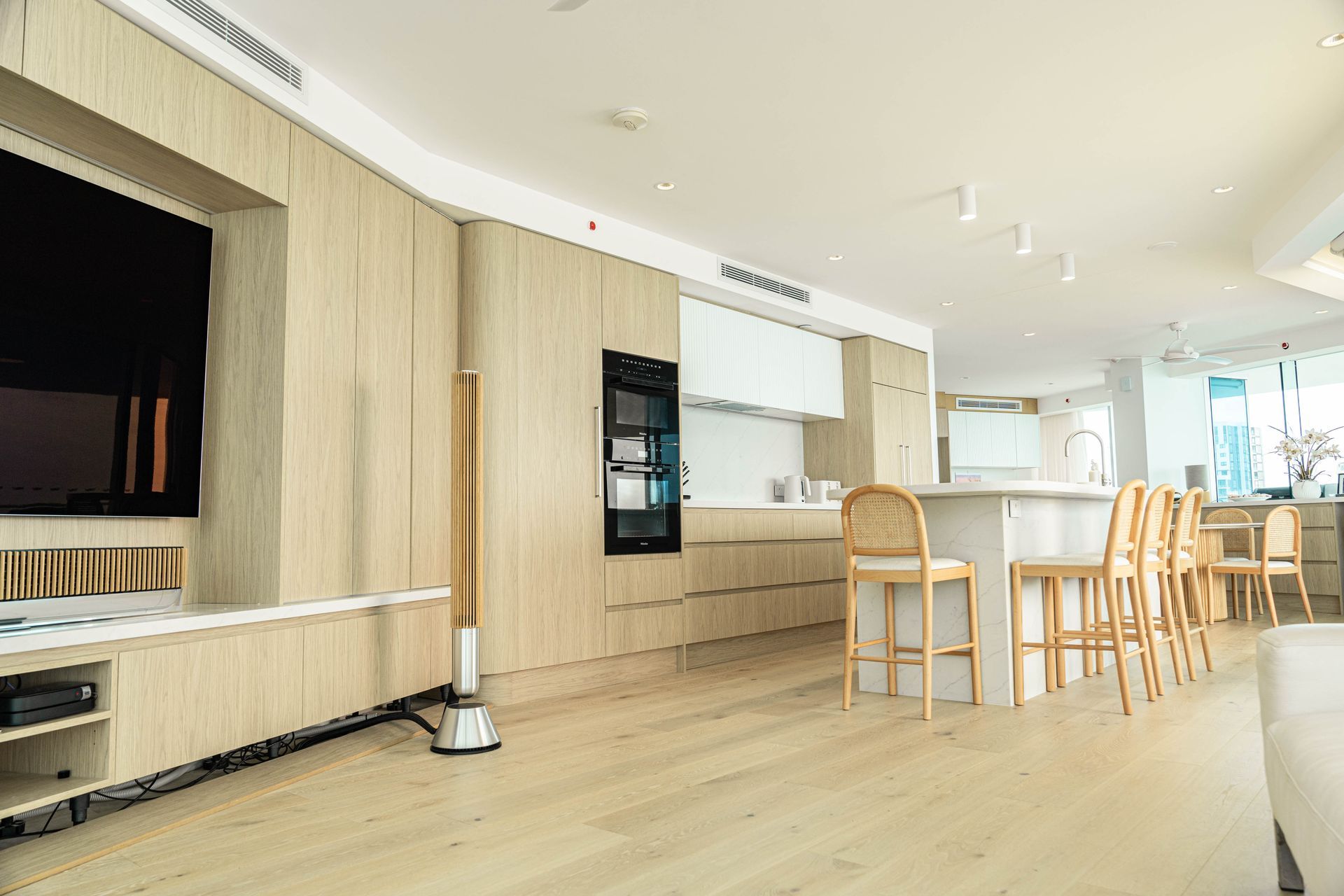 A modern, open-plan kitchen and living room featuring light wood cabinetry, white countertops, and a dining island.