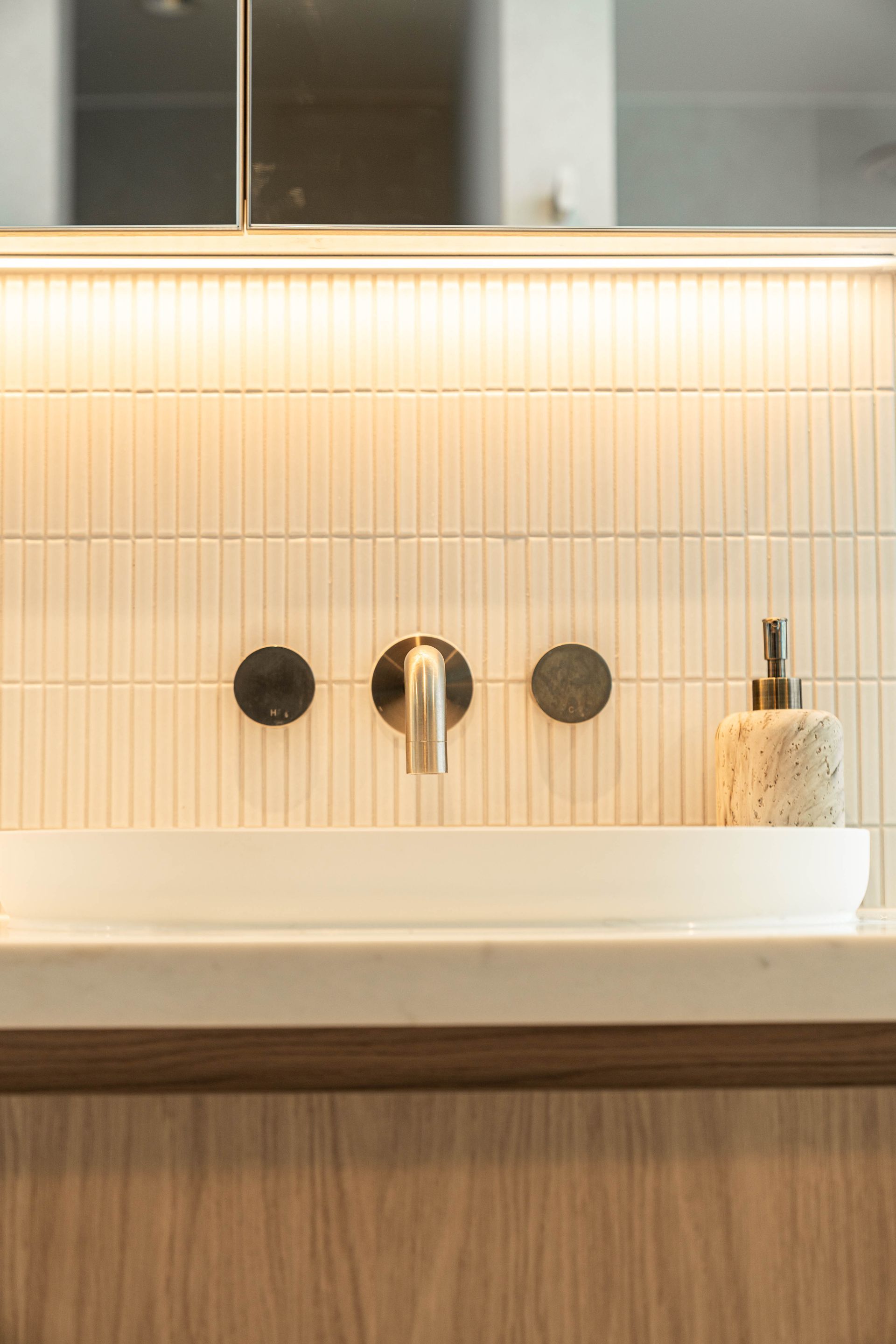 A bathroom vanity with white vertical tiles, a round white sink, wall-mounted brass faucet, and a stone soap dispenser.