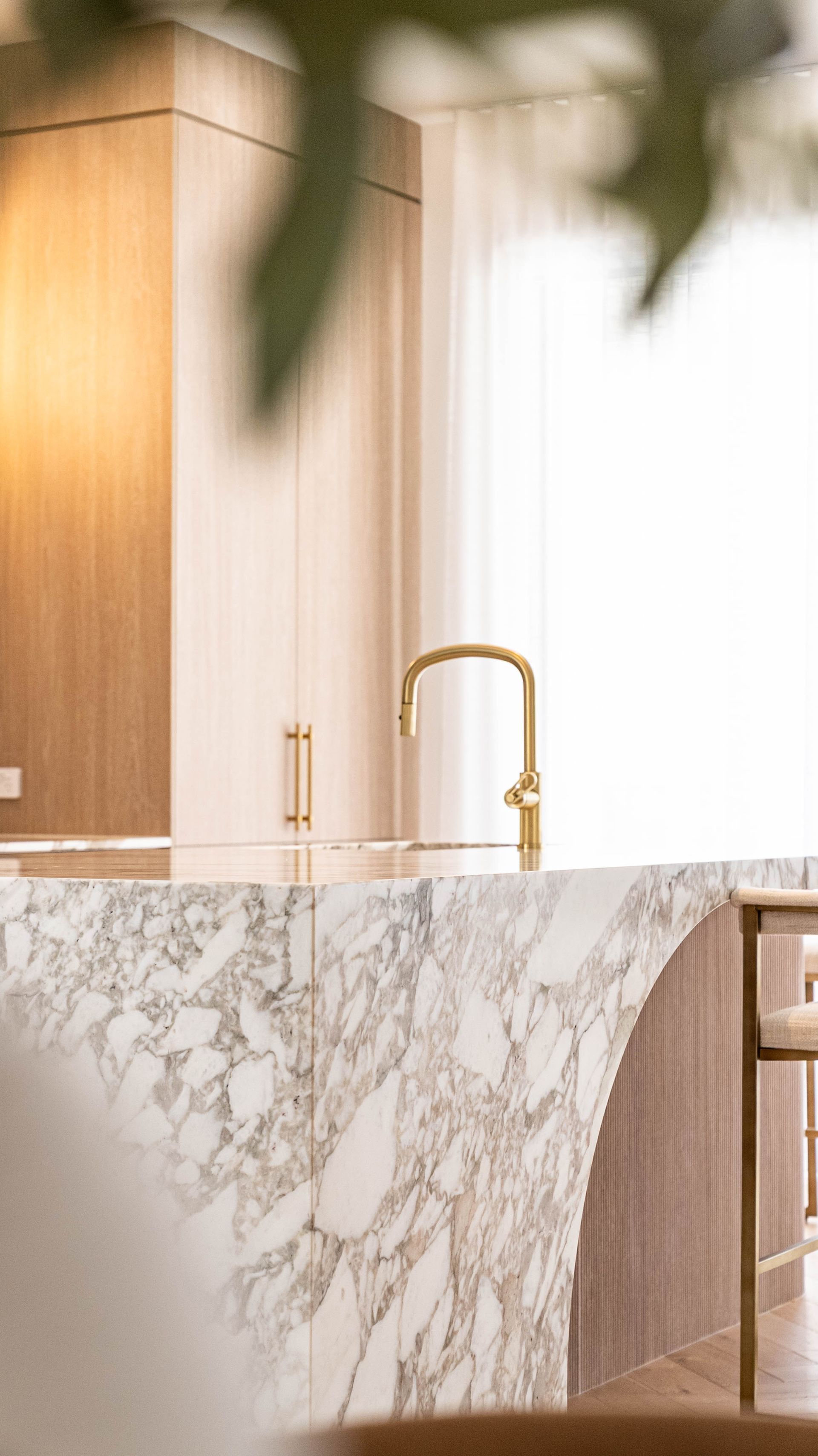 A modern kitchen island with a marble countertop, gold faucet, and light wood cabinetry, viewed through a foreground plant.