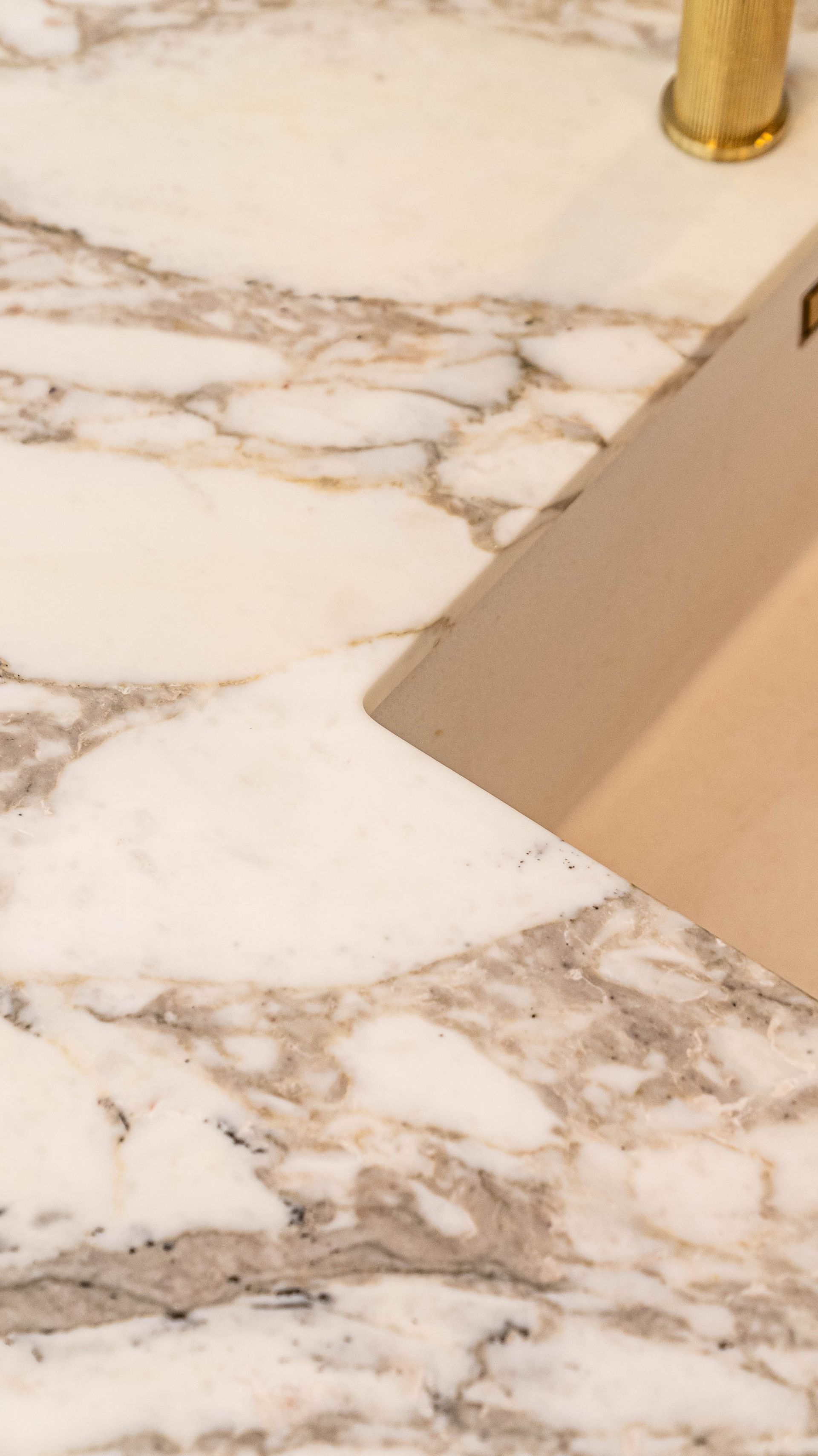 A metal putty knife scrapes a smooth, tan paste across a white and grey marble countertop.