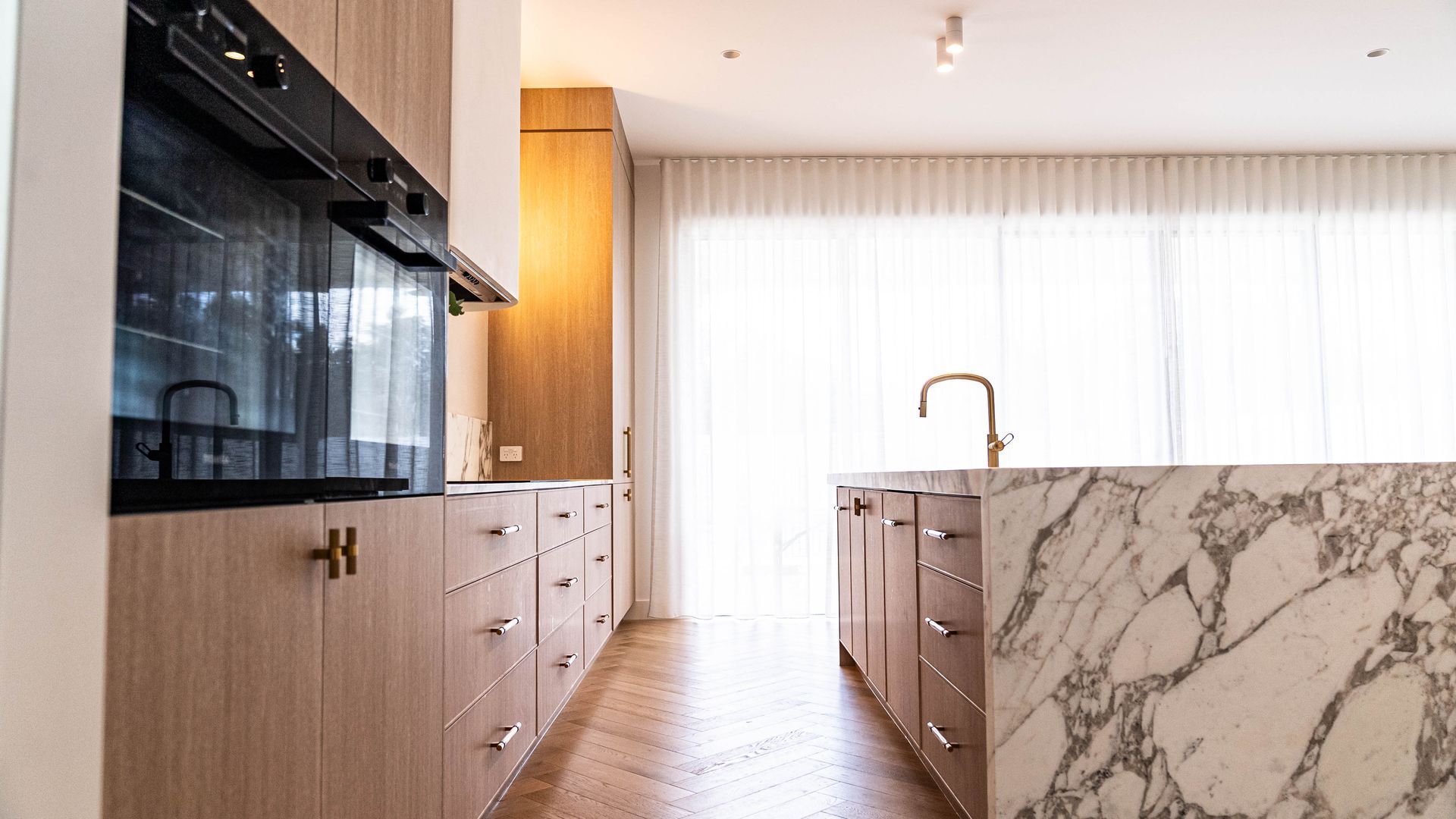 A modern kitchen with light wood cabinetry, a marble-topped island, and herringbone wood floors.
