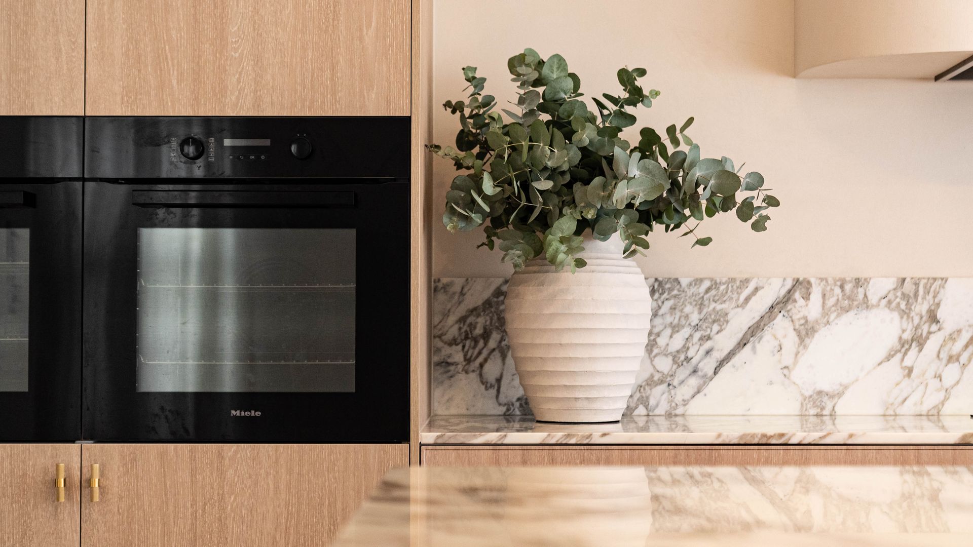 A modern kitchen with a built-in black oven set in light wood cabinetry, featuring a marble countertop and vase of leaves.