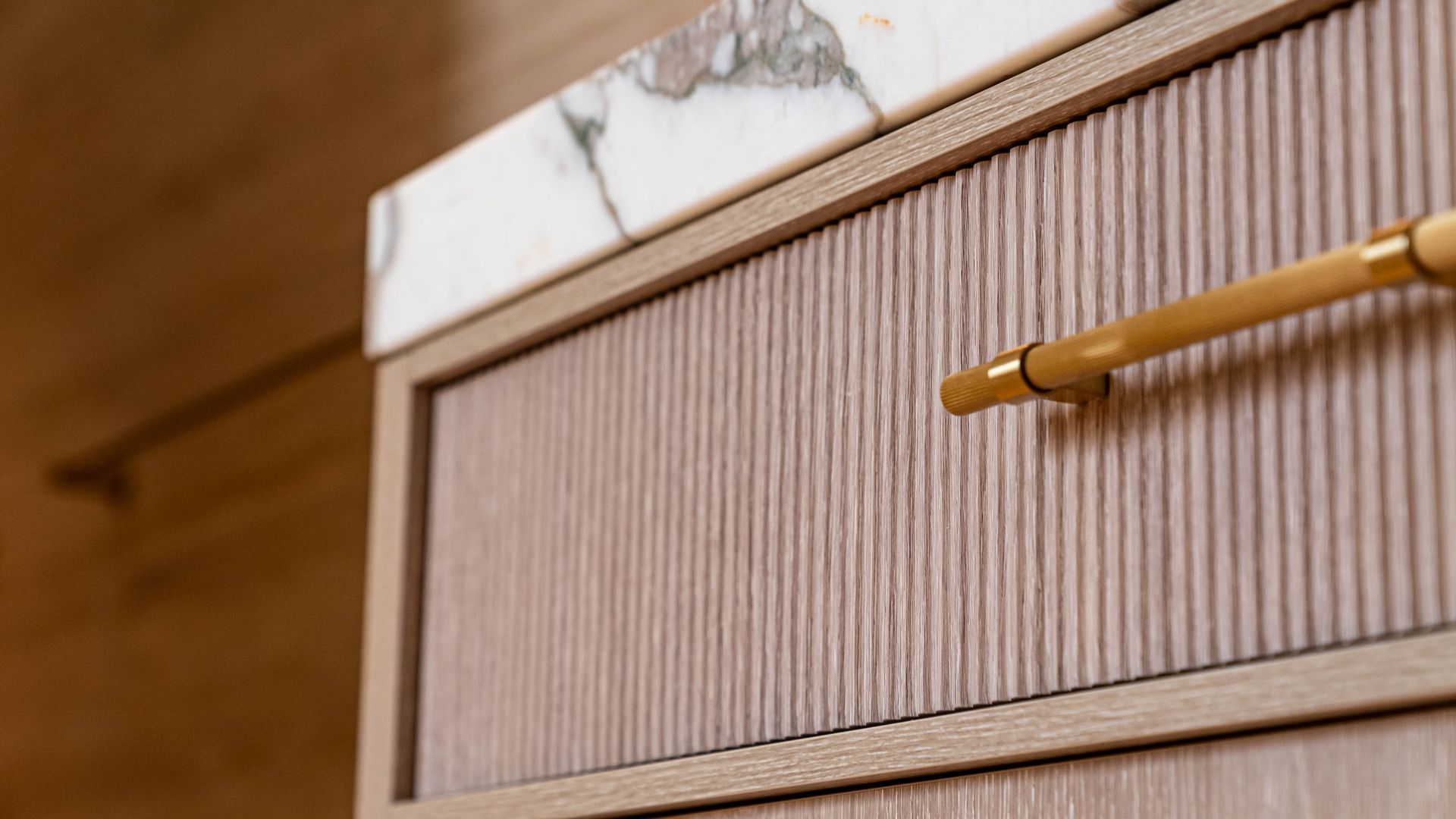 A close-up of a wooden vanity drawer with vertical reeding, a marble countertop, and a sleek gold handle.