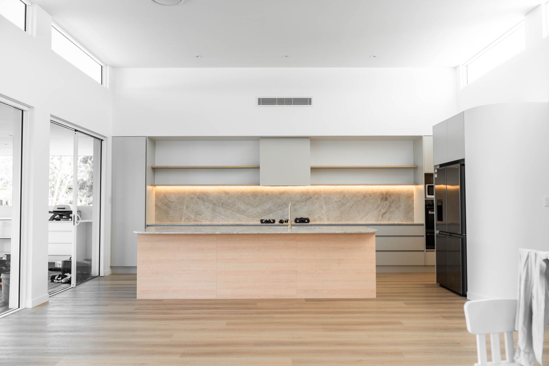 Minimalist kitchen with white walls, light timber flooring and a central island finished with a custom stone benchtop in a Gold Coast home, complemented by a marble splashback and open shelving